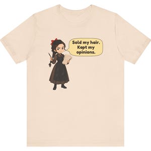 May include: A light beige t-shirt featuring a cartoon illustration of a person with dark hair, a red bow, and a brown dress. The character holds a lock of hair and a paper, with a speech bubble containing the text: Sold my hair. Kept my opinions.