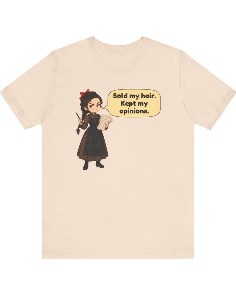 May include: A light beige t-shirt featuring a cartoon illustration of a person with dark hair, a red bow, and a brown dress. The character holds a lock of hair and a paper, with a speech bubble containing the text: Sold my hair. Kept my opinions.