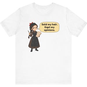 May include: A white t-shirt featuring a cartoon illustration of a person with dark braided hair, wearing a brown dress and black apron, holding a quill and paper. A speech bubble contains the text: Sold my hair. Kept my opinions.
