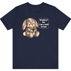 May include: A navy blue short-sleeve t-shirt featuring a graphic of a brown stuffed rabbit holding holly berries. A speech bubble next to the rabbit contains the text: Shabby? No. Very well loved. The rabbit has a white tail and stitched details.