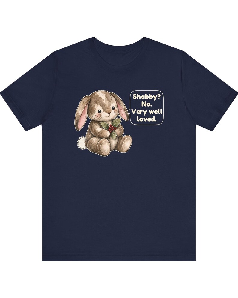 May include: A navy blue short-sleeve t-shirt featuring a graphic of a brown stuffed rabbit holding holly berries. A speech bubble next to the rabbit contains the text: Shabby? No. Very well loved. The rabbit has a white tail and stitched details.
