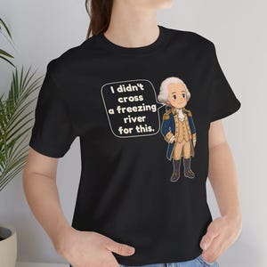 May include: A black t-shirt featuring a cartoon illustration of a historical figure in a blue and tan military uniform. Next to the figure is a white speech bubble containing the text, I didn't cross a freezing river for this.