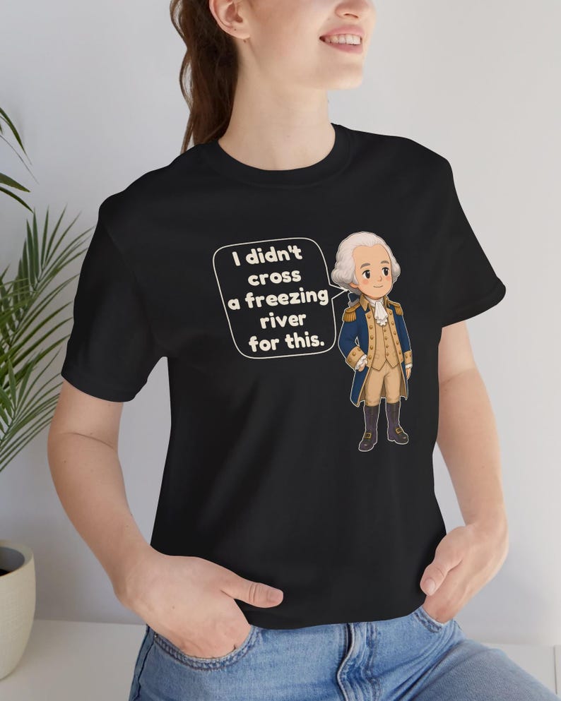 May include: A black t-shirt featuring a cartoon illustration of a historical figure in a blue and tan military uniform. Next to the figure is a white speech bubble containing the text, I didn't cross a freezing river for this.