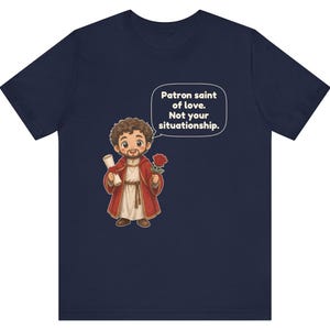 Puede incluir: Una camiseta azul marino con una ilustraci&oacute;n de dibujos animados de una figura barbuda con t&uacute;nica roja que sostiene un pergamino y una rosa roja. Un globo de texto sobre la figura contiene el texto: Patron saint of love. Not your situationship.