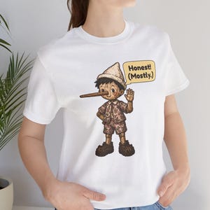 K&ouml;nnte beinhalten: Ein wei&szlig;es Kurzarm-T-Shirt mit einer grafischen Illustration einer Holzpuppe mit langer Nase, die einen Hut und ein gemustertes Outfit tr&auml;gt. Eine gelbe Sprechblase neben der Figur enth&auml;lt den Text Honest! (Mostly.)