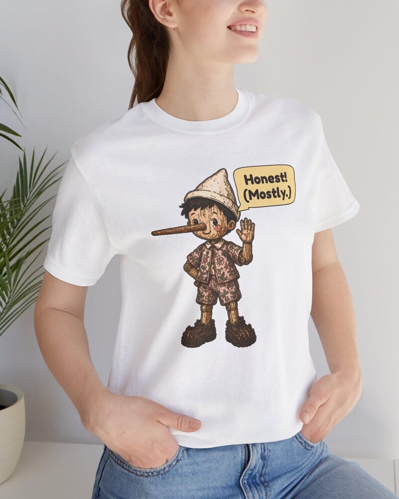 K&ouml;nnte beinhalten: Ein wei&szlig;es Kurzarm-T-Shirt mit einer grafischen Illustration einer Holzpuppe mit langer Nase, die einen Hut und ein gemustertes Outfit tr&auml;gt. Eine gelbe Sprechblase neben der Figur enth&auml;lt den Text Honest! (Mostly.)