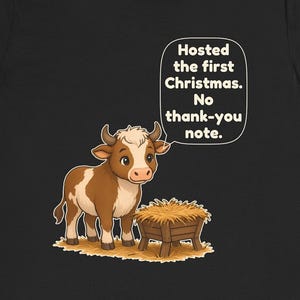 以下が含まれることがあります： 黒いTシャツに、干し草が入った木製の飼い葉桶の横に立つ茶色と白の子牛が描かれています。子牛の上の吹き出しには「Hosted the first Christmas. No thank-you note.」というテキストが含まれています。