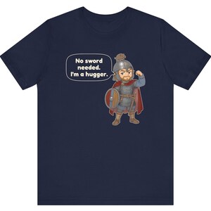 May include: A navy blue t-shirt featuring a cartoon illustration of a warrior in chainmail and a red cape, wearing a helmet with a small animal on top. The warrior flexes an arm and a speech bubble contains the text: No sword needed. I'm a hugger.