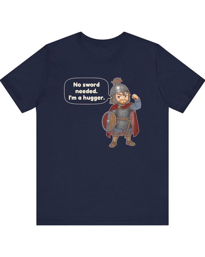 May include: A navy blue t-shirt featuring a cartoon illustration of a warrior in chainmail and a red cape, wearing a helmet with a small animal on top. The warrior flexes an arm and a speech bubble contains the text: No sword needed. I'm a hugger.