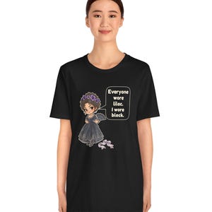 May include: A black t-shirt featuring a graphic of a cartoon character in a dark dress with a purple flower crown and a fan. A speech bubble contains the text: Everyone wore lilac. I wore black. A small purple ribbon is positioned below the character.