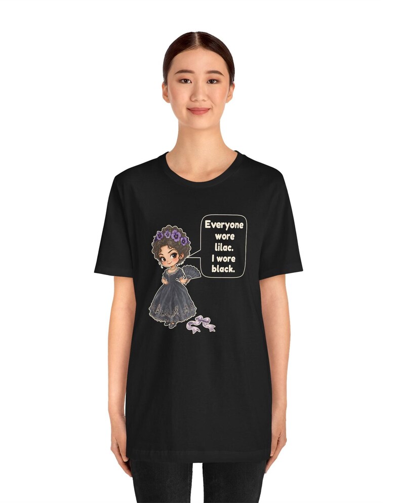 May include: A black t-shirt featuring a graphic of a cartoon character in a dark dress with a purple flower crown and a fan. A speech bubble contains the text: Everyone wore lilac. I wore black. A small purple ribbon is positioned below the character.