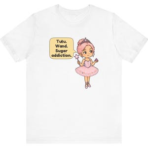 May include: A white t-shirt featuring a cartoon illustration of a ballerina with pink hair, a tiara, and a pink tutu. The character holds a star-tipped wand and a small treat. A speech bubble contains the text: Tutu. Wand. Sugar addiction.