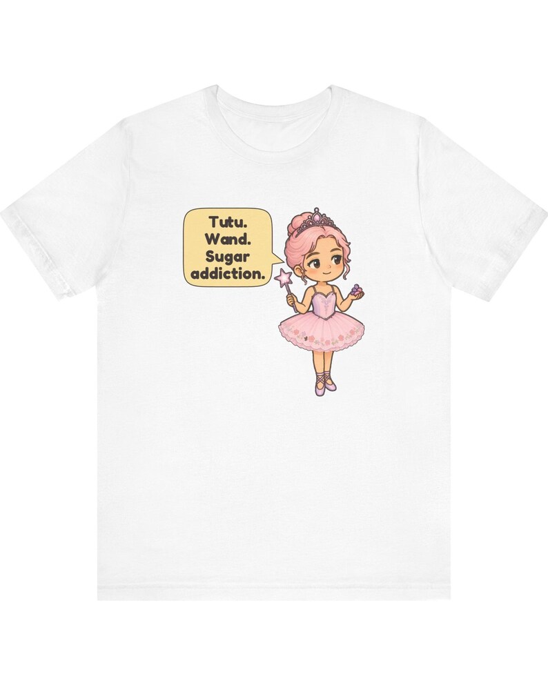 May include: A white t-shirt featuring a cartoon illustration of a ballerina with pink hair, a tiara, and a pink tutu. The character holds a star-tipped wand and a small treat. A speech bubble contains the text: Tutu. Wand. Sugar addiction.