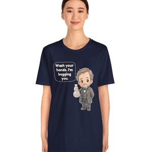 May include: A navy blue short-sleeve t-shirt featuring a cartoon illustration of a man in a suit holding a glass flask. A speech bubble next to the character contains the text: Wash your hands. I'm begging you. The design is centered on the chest.