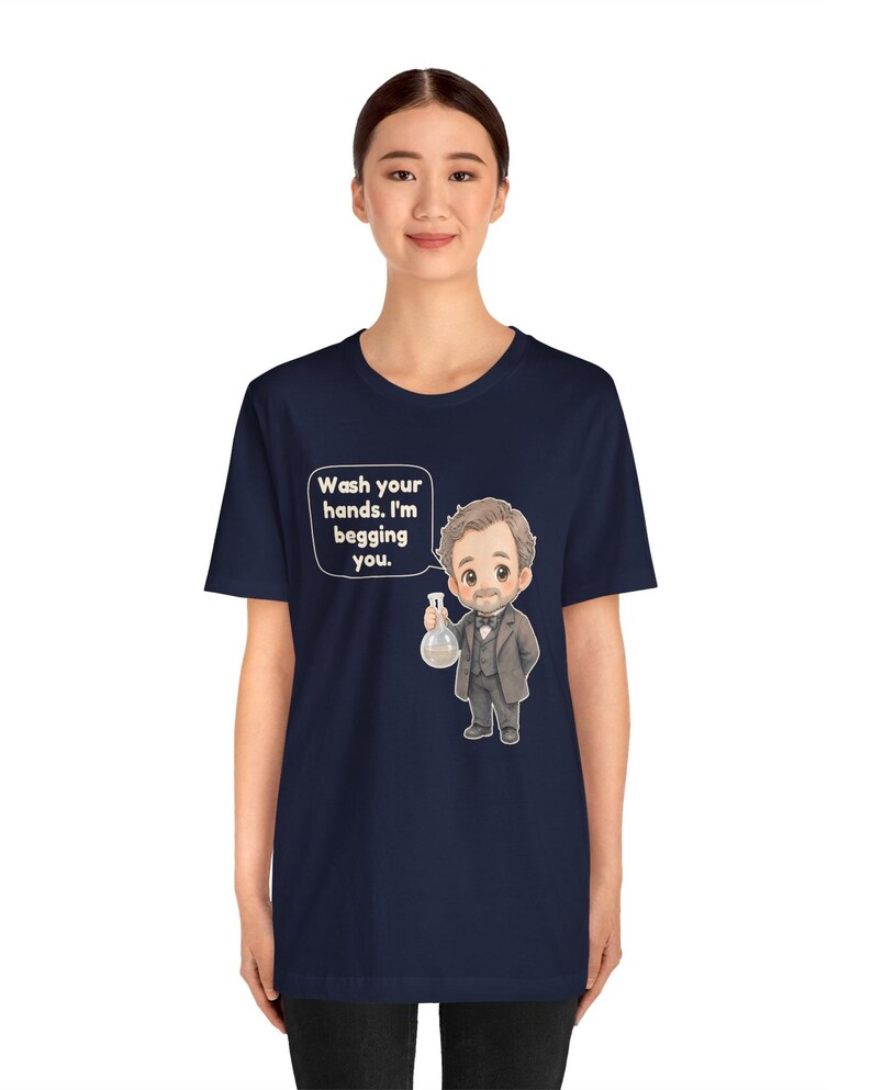 May include: A navy blue short-sleeve t-shirt featuring a cartoon illustration of a man in a suit holding a glass flask. A speech bubble next to the character contains the text: Wash your hands. I'm begging you. The design is centered on the chest.