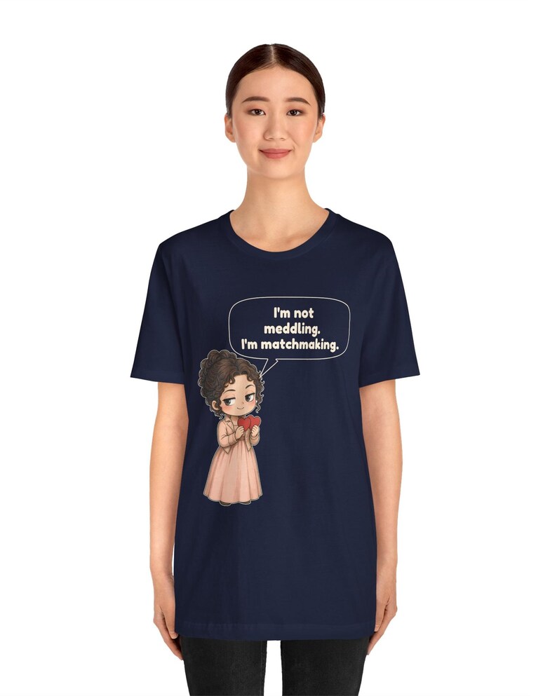 K&ouml;nnte beinhalten: Ein kurz&auml;rmeliges, marineblaues T-Shirt mit einer Cartoon-Illustration einer Person mit braunen Haaren, die ein hellrosa Kleid und eine Jacke tr&auml;gt und ein rotes Herz h&auml;lt. Eine Sprechblase dar&uuml;ber enth&auml;lt den Text: I'm not meddling. I'm matchmaking.
