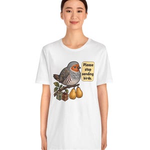 K&ouml;nnte beinhalten: Ein wei&szlig;es T-Shirt mit einer Grafik eines grauen und orangefarbenen Vogels, der auf einem Ast mit zwei Birnen und zwei kleinen Geschenkkartons sitzt. Eine Sprechblase neben dem Vogel enth&auml;lt den Text: "Please stop sending birds."