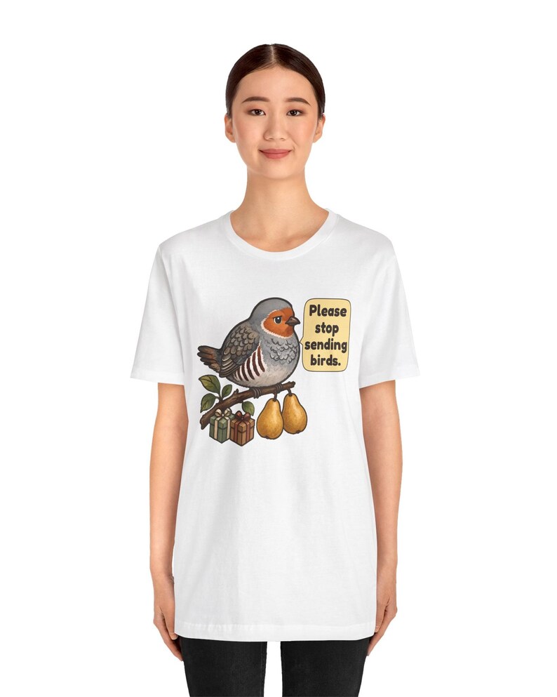 K&ouml;nnte beinhalten: Ein wei&szlig;es T-Shirt mit einer Grafik eines grauen und orangefarbenen Vogels, der auf einem Ast mit zwei Birnen und zwei kleinen Geschenkkartons sitzt. Eine Sprechblase neben dem Vogel enth&auml;lt den Text: "Please stop sending birds."