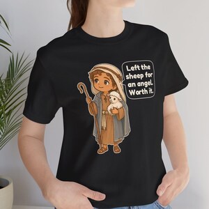 May include: A black t-shirt featuring a cartoon illustration of a shepherd holding a small white lamb and a wooden staff. The shepherd wears a tan tunic and a blue cloak. A speech bubble contains the text: Left the sheep for an angel. Worth it.