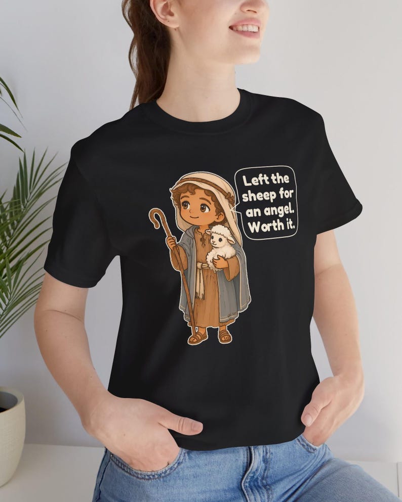 May include: A black t-shirt featuring a cartoon illustration of a shepherd holding a small white lamb and a wooden staff. The shepherd wears a tan tunic and a blue cloak. A speech bubble contains the text: Left the sheep for an angel. Worth it.