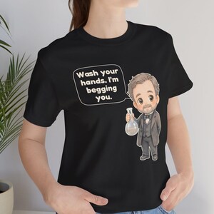 May include: A black t-shirt featuring a cartoon illustration of a man in a suit holding a glass flask. A speech bubble above the figure contains the text: Wash your hands. I'm begging you. The design is centered on the front of the garment.