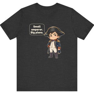 May include: A dark heather gray t-shirt featuring a cartoon illustration of a small figure dressed in a blue and white military-style uniform with a bicorne hat. A speech bubble next to the character contains the text: Small emperor. Big plans.