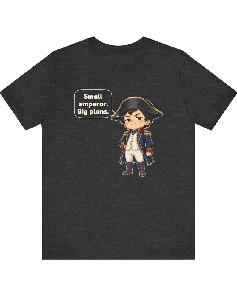May include: A dark heather gray t-shirt featuring a cartoon illustration of a small figure dressed in a blue and white military-style uniform with a bicorne hat. A speech bubble next to the character contains the text: Small emperor. Big plans.