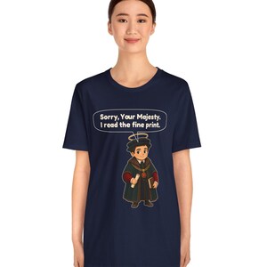 May include: A navy blue t-shirt featuring a graphic of a cartoon character wearing a dark robe, a halo, and holding a scroll and book. Above the character is a speech bubble with the white text: Sorry, Your Majesty. I read the fine print.