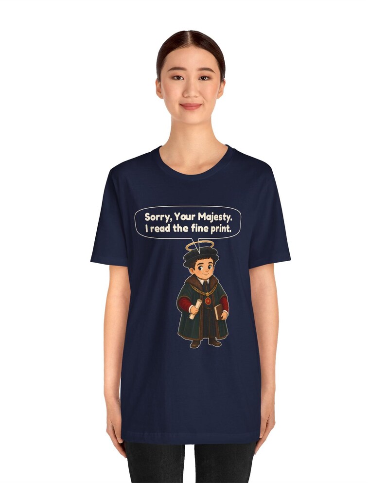 May include: A navy blue t-shirt featuring a graphic of a cartoon character wearing a dark robe, a halo, and holding a scroll and book. Above the character is a speech bubble with the white text: Sorry, Your Majesty. I read the fine print.