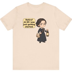 May include: A light beige t-shirt featuring a cartoon illustration of a person in a dark dress, carrying a satchel and a lit lantern. A speech bubble contains the text: Retire? At 59 I was just getting started.