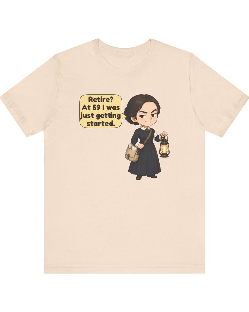May include: A light beige t-shirt featuring a cartoon illustration of a person in a dark dress, carrying a satchel and a lit lantern. A speech bubble contains the text: Retire? At 59 I was just getting started.