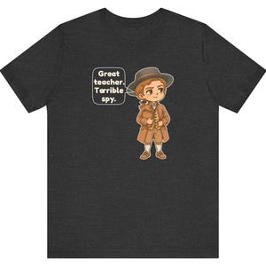 May include: A dark heather gray t-shirt featuring a cartoon illustration of a person in 18th-century brown clothing and a hat, holding a book. A speech bubble next to the character contains the text: Great teacher. Terrible spy.