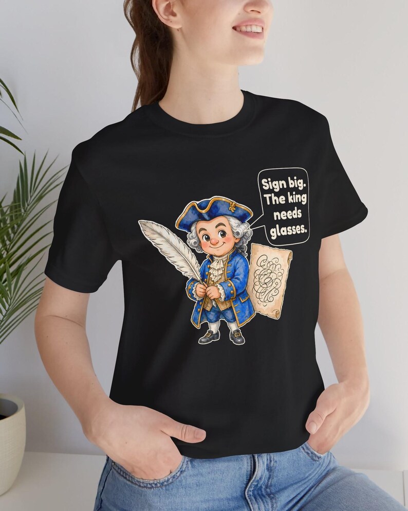May include: A black t-shirt featuring a cartoon illustration of a person in an 18th-century blue coat and tricorn hat holding a large white quill and a scroll. A speech bubble contains the text: Sign big. The king needs glasses.