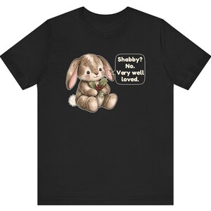 May include: A black short-sleeved t-shirt featuring a graphic of a brown stuffed rabbit holding a sprig of holly with red berries. Next to the rabbit is a white speech bubble containing the text: Shabby? No. Very well loved.
