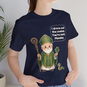 Puede incluir: Una camiseta azul marino con una ilustraci&oacute;n de dibujos animados de San Patricio sosteniendo un bast&oacute;n de madera y un tr&eacute;bol. Una peque&ntilde;a serpiente verde est&aacute; a sus pies. Un bocadillo contiene el texto: I drove out the snakes. You're next, Monday.