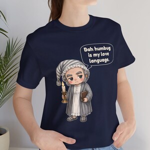 Puede incluir: Una camiseta azul marino con el gr&aacute;fico de un personaje con gorro de dormir y bata a rayas, sosteniendo una vela encendida y un libro. Un globo de texto contiene la frase: Bah humbug is my love language.