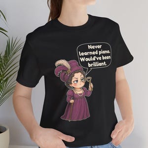 May include: A black t-shirt featuring a graphic of a person in a purple dress and hat, holding a monocle and a cane. A speech bubble above the figure contains the text: Never learned piano. Would've been brilliant.