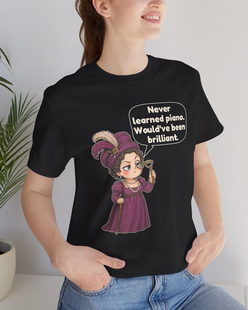 May include: A black t-shirt featuring a graphic of a person in a purple dress and hat, holding a monocle and a cane. A speech bubble above the figure contains the text: Never learned piano. Would've been brilliant.