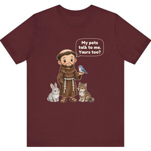 May include: A maroon short-sleeve t-shirt featuring a cartoon illustration of a monk in a brown robe with a rabbit, a wolf, and a blue bird. A speech bubble above the monk contains the text: My pets talk to me. Yours too?