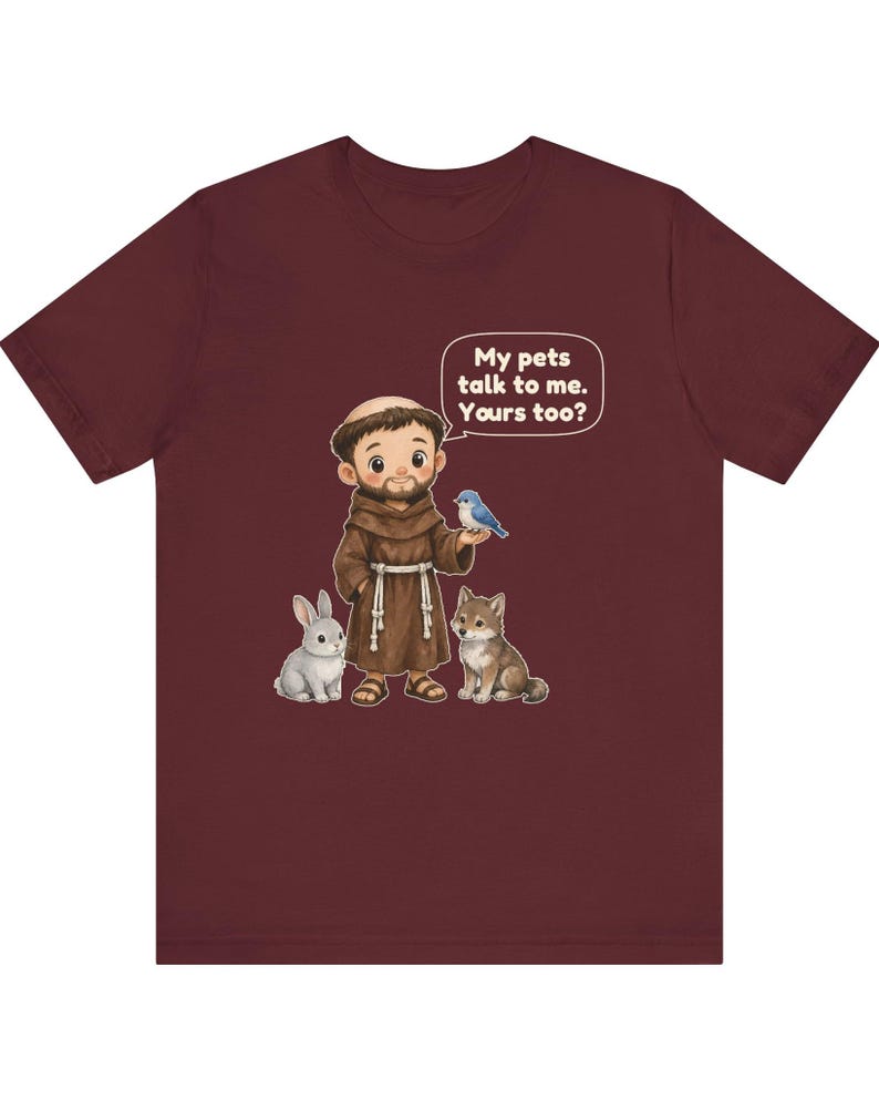 May include: A maroon short-sleeve t-shirt featuring a cartoon illustration of a monk in a brown robe with a rabbit, a wolf, and a blue bird. A speech bubble above the monk contains the text: My pets talk to me. Yours too?