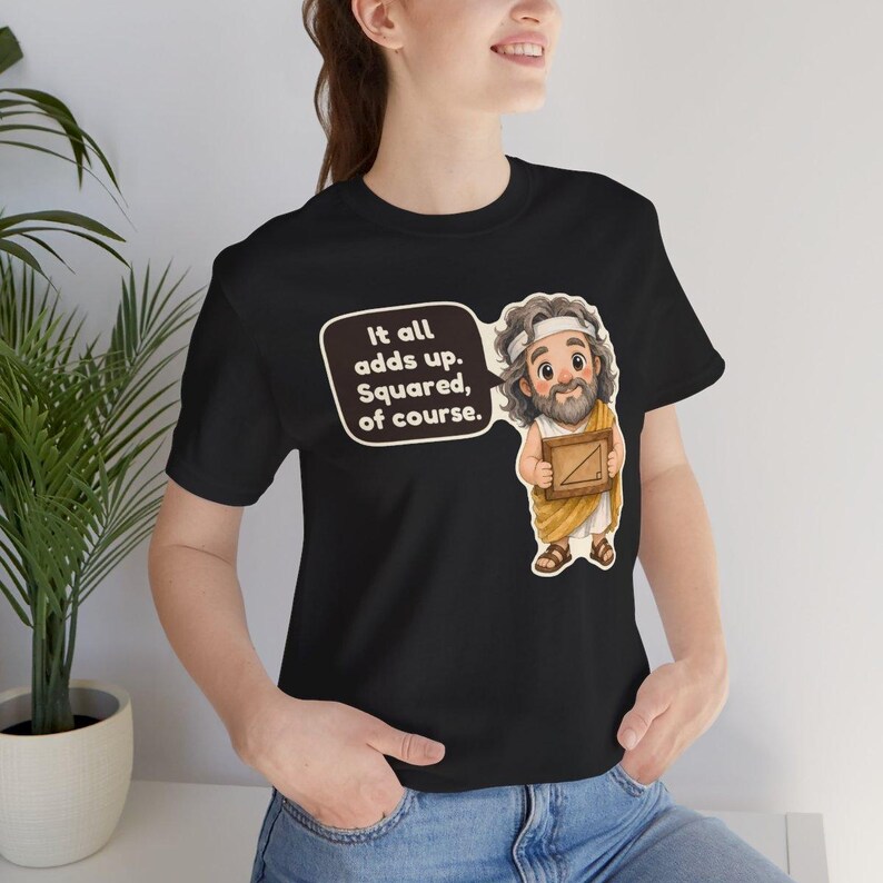 Pu&ograve; includere: Una t-shirt nera con un'illustrazione a cartone animato di un uomo barbuto in toga gialla che tiene una cornice di legno con un triangolo. Un fumetto accanto contiene il testo: It all adds up. Squared, of course.