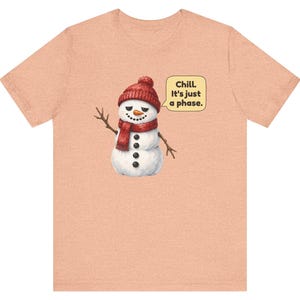 Pode incluir: Uma t-shirt cor de p&ecirc;ssego com o gr&aacute;fico de um boneco de neve a usar um gorro e um cachecol de malha vermelhos. O boneco de neve tem um nariz de cenoura, bra&ccedil;os de pau e um bal&atilde;o com o texto: Chill. It's just a phase.