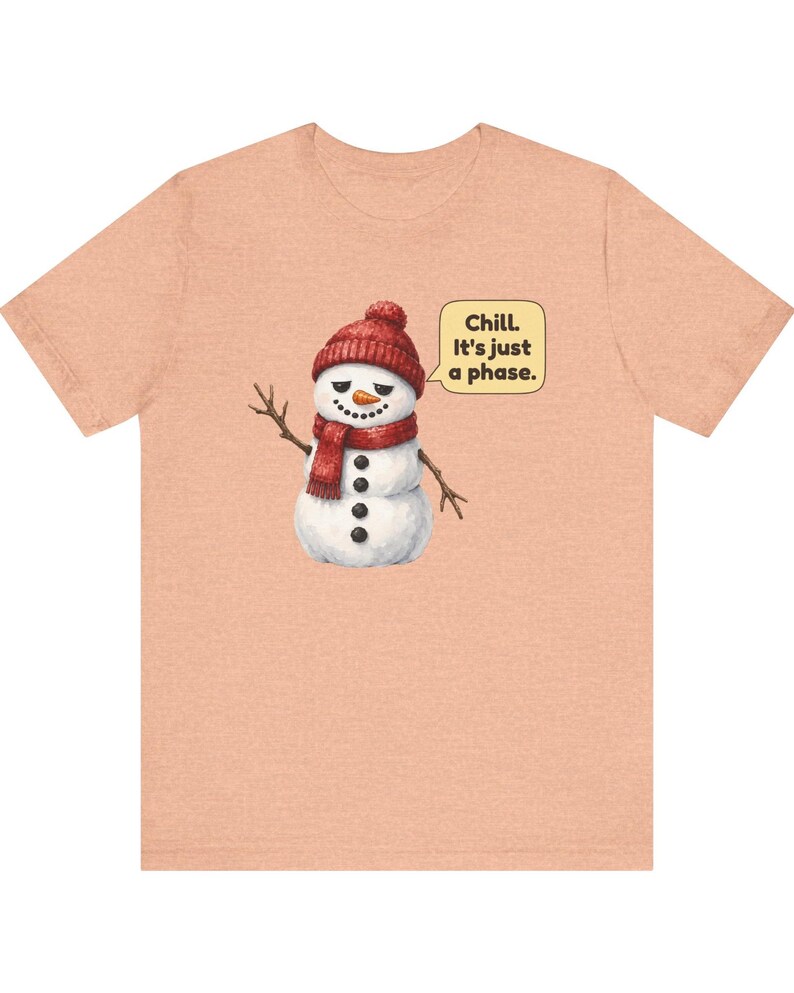 Pode incluir: Uma t-shirt cor de p&ecirc;ssego com o gr&aacute;fico de um boneco de neve a usar um gorro e um cachecol de malha vermelhos. O boneco de neve tem um nariz de cenoura, bra&ccedil;os de pau e um bal&atilde;o com o texto: Chill. It's just a phase.