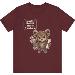 Puede incluir: Una camiseta de manga corta color granate con el gr&aacute;fico de una peque&ntilde;a criatura peluda con cuernos que sostiene un manojo de palos y un pergamino. Un bocadillo sobre la criatura contiene el texto: Naughty list? I'll take it from here.