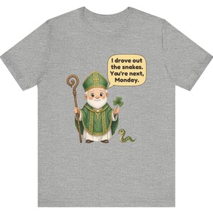 Puede incluir: Una camiseta gris jaspeado con una ilustraci&oacute;n de dibujos animados de una figura barbuda con vestimentas religiosas verdes que sostiene un bast&oacute;n de madera y un tr&eacute;bol. Una peque&ntilde;a serpiente verde est&aacute; cerca y un globo de texto dice: I drove out the snakes. You're next, Monday.