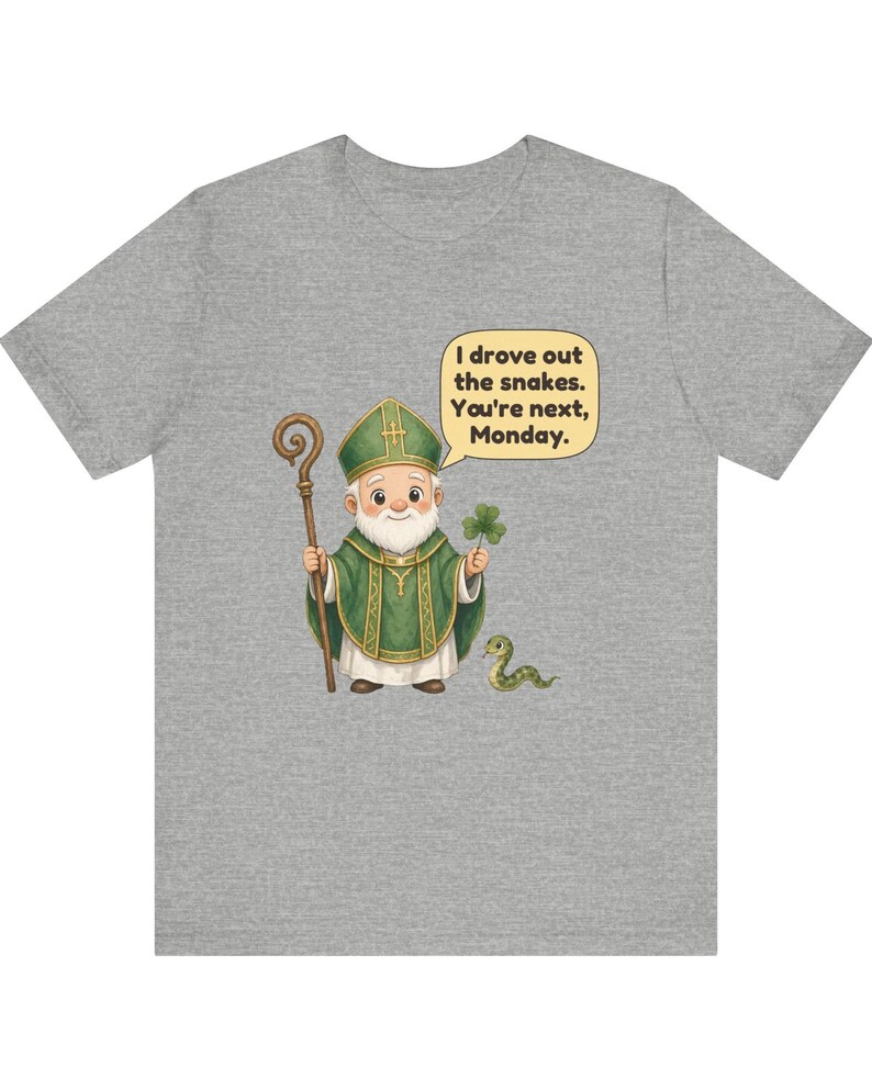 Puede incluir: Una camiseta gris jaspeado con una ilustraci&oacute;n de dibujos animados de una figura barbuda con vestimentas religiosas verdes que sostiene un bast&oacute;n de madera y un tr&eacute;bol. Una peque&ntilde;a serpiente verde est&aacute; cerca y un globo de texto dice: I drove out the snakes. You're next, Monday.