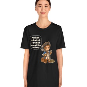 May include: A black t-shirt featuring a cartoon illustration of a historical figure in a blue coat and tricorn hat holding a shovel and compass, with a speech bubble containing the text: Arrived uninvited. Fortified everything anyway.