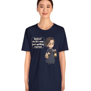 May include: A navy blue short-sleeve t-shirt featuring a cartoon illustration of a person in a dark dress holding a lantern and a satchel. A speech bubble next to the character contains the text: Retire? At 59 I was just getting started.
