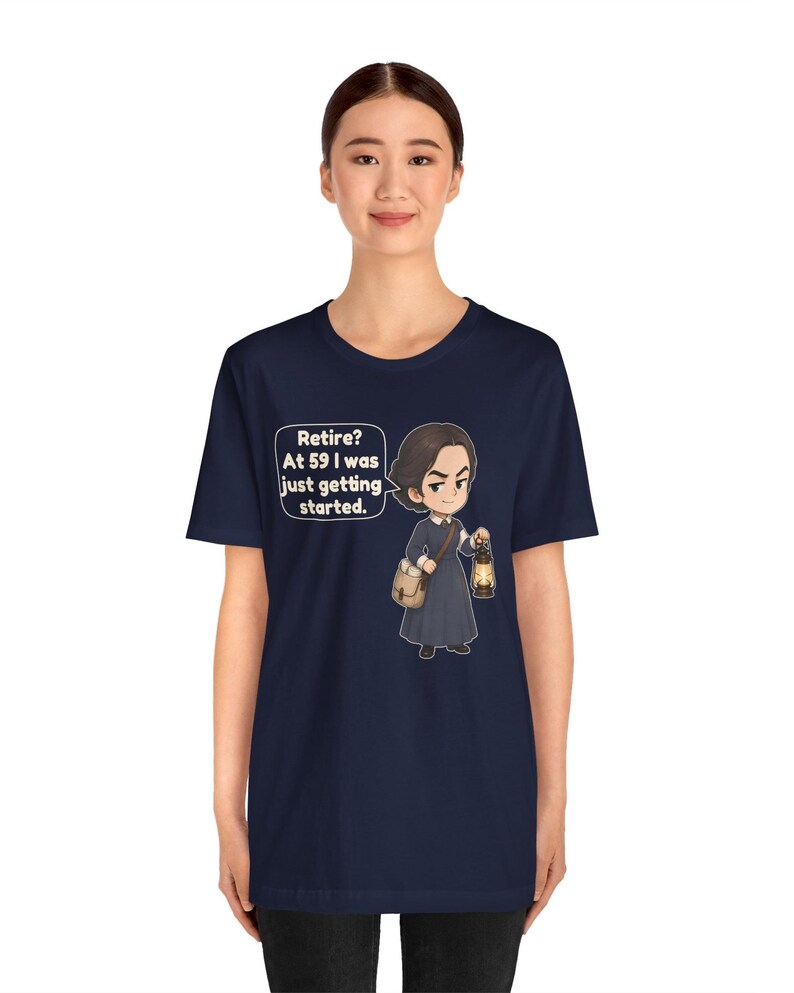 May include: A navy blue short-sleeve t-shirt featuring a cartoon illustration of a person in a dark dress holding a lantern and a satchel. A speech bubble next to the character contains the text: Retire? At 59 I was just getting started.