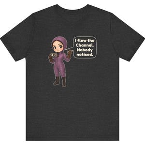 Puede incluir: Una camiseta gris oscuro con una ilustraci&oacute;n de dibujos animados de una persona con traje de vuelo morado y gafas. Un globo de texto junto al personaje contiene el texto: I flew the Channel. Nobody noticed.